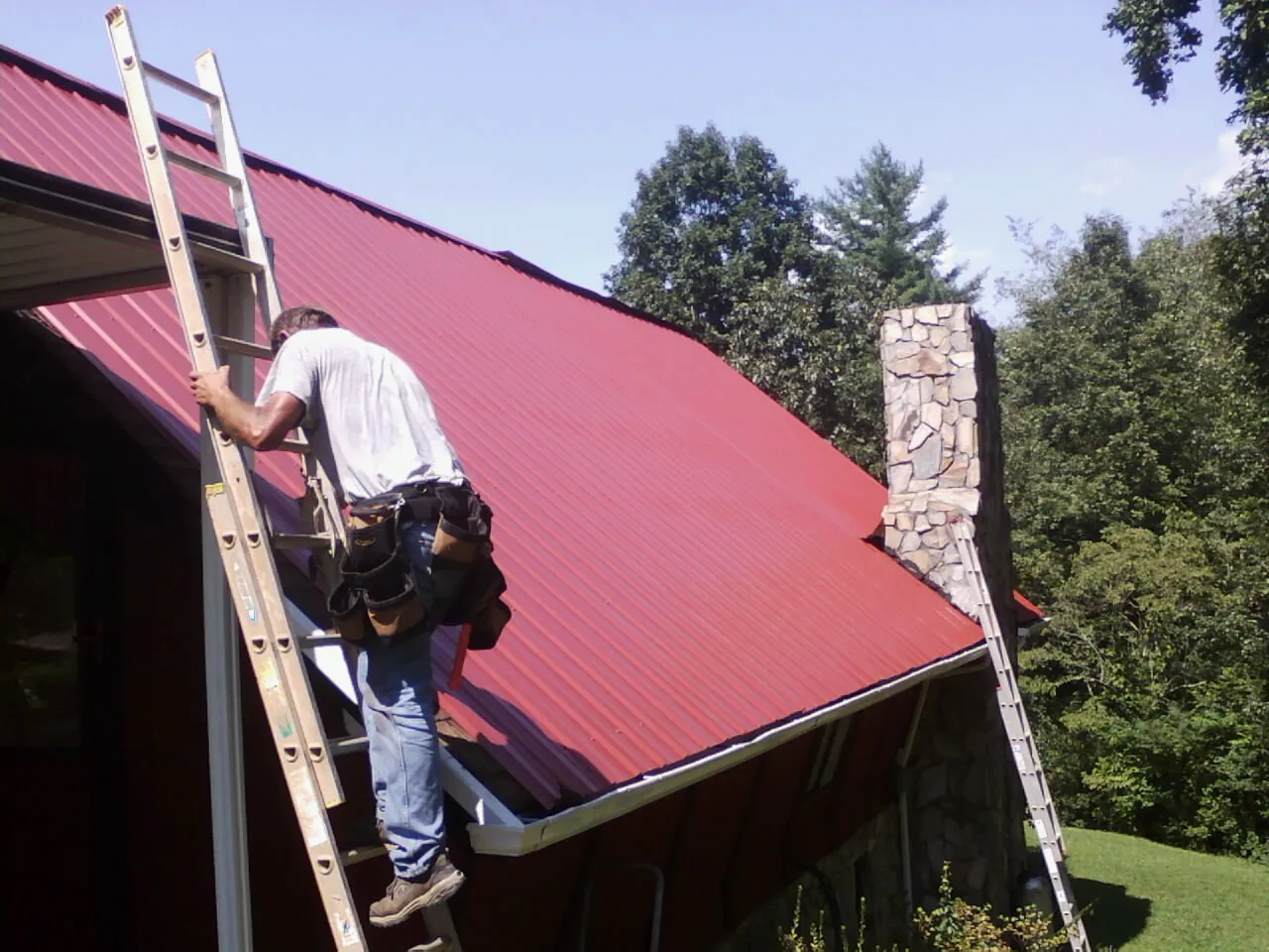 Roofer on a ladder beside a red metal roof and stone chimney — Soffit & Fascia Repair by Roofing Brothers in Exeter